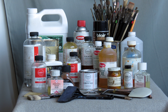 photo of painting materials on a table including bottles of solvent and mediums, brushes, varnishes, and palette knives